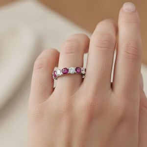 Puede incluir: Un anillo de plata con piedras preciosas rojas y claras alternas. El diseño del anillo presenta una serie de piedras rojas redondas y vibrantes intercaladas con piedras claras y brillantes. El anillo está en una mano, destacando su elegante diseño.
