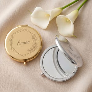 May include: Two round compact mirrors, one gold and one silver, are displayed. The gold mirror is closed and features the name "Emma" inside a geometric design with floral accents. The silver mirror is open, revealing a reflective surface.