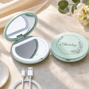 May include: A round, mint green compact mirror with LED lighting. One mirror is open, showing a circular light ring and a power button icon. The other mirror is closed, featuring a black line-art floral frame and the name Shirley printed in script.