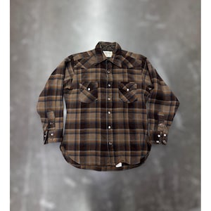 Puede incluir: Una camisa de manga larga con botones y un patrón de cuadros en tonos marrón, negro y beige. La prenda incluye dos bolsillos en el pecho con cierres de presión, un cuello puntiagudo y botones de presión en la parte frontal y en los puños. El forro interior del cuello tiene una textura acolchada.