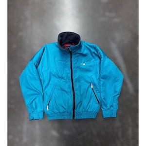 Puede incluir: Una chaqueta con cremallera de color turquesa brillante que presenta un cuello alto con forro polar gris oscuro, una cremallera frontal negra completa, dos bolsillos laterales con cremallera y un pequeño logo blanco bordado en el pecho.