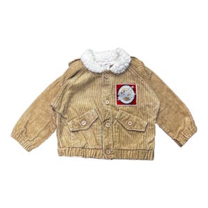 Puede incluir: Una chaqueta de pana color canela con cuello de sherpa blanco, mangas largas con puños elásticos y cierre de botones. La chaqueta incluye dos bolsillos delanteros con solapa y botones, y un parche cuadrado en el pecho con un avión.
