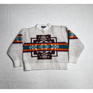 Può includere: Un maglione a maniche lunghe con girocollo in maglia grigio chiaro mélange. La parte anteriore presenta un grande motivo geometrico marrone scuro centrato orizzontalmente con una fascia vivace di strisce color ottanio, arancione e rosso.