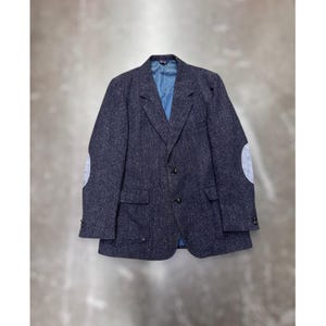 Puede incluir: Un blazer gris oscuro con un patrón de espiga, con coderas grises claras y un forro interior azul. La chaqueta tiene dos bolsillos delanteros y un cierre de dos botones. Una prenda clásica y a medida.