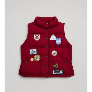 May include: A deep red puffer vest featuring a high collar and a front snap-button closure. The garment is decorated with various embroidered patches, including text that reads Taos, Aspen, Snow, and Polo Jeans Company, along with mountain motifs.