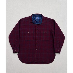 Puede incluir: Una camisa de manga larga con botones en color burdeos intenso con un patrón de cuadrícula azul oscuro. La camisa presenta dos bolsillos en el pecho, un cuello en punta y un forro acolchado azul oscuro visible en el cuello. Puños y tapeta con botones.