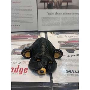 Puede incluir: Una figura de oso negro con detalles de pelaje texturizado, que presenta acentos de color beige en la nariz, las orejas y las almohadillas de sus cuatro patas. El oso está representado en una posición tumbada con la cabeza entre las patas.