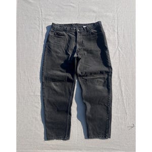 Puede incluir: Unos pantalones vaqueros de color negro carbón con un diseño clásico de cinco bolsillos, cierre de botones, trabillas para el cinturón en la cintura y costuras tonales visibles en toda la tela, presentados en un corte recto estándar.