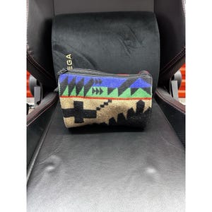 Puede incluir: Un estuche rectangular con cremallera que presenta un patrón geométrico multicolor con bandas en azul, verde, beige y negro. El diseño incluye motivos triangulares y en forma de cruz sobre la superficie de tela y cierre de cremallera negro.
