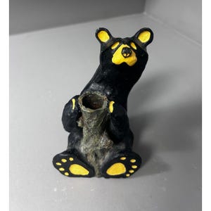 Pode incluir: Uma estatueta de urso preto com detalhes amarelos nas orelhas, focinho e patas. O urso está sentado, segurando um objeto cilíndrico texturizado cinza-acastanhado que apresenta uma abertura circular oca no topo da estrutura.