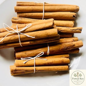 May include: A collection of light brown, cylindrical cinnamon sticks arranged in bundles and tied with thin white string. The sticks have a rough, woody texture and are stacked together. A small square logo with the text Forest Key is visible.