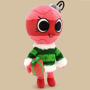 Könnte beinhalten: Ein Plüsch-Weihnachtsschmuck-Charakter mit einem lächelnden Gesicht, der einen grünen Pullover mit weißem Besatz trägt und ein rot-grünes Geschenk hält. Der Schmuck ist rot und rosa mit schwarzen Augen und einer silbernen Kappe.