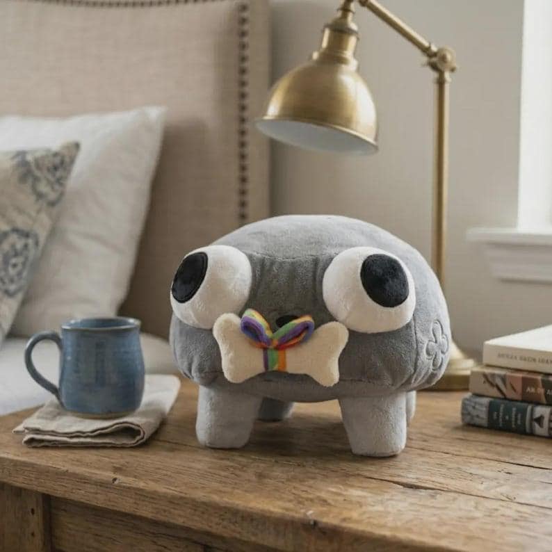 May include: A gray plush toy with large white eyes and black pupils, a bone-shaped bow tie with rainbow ribbon, and four small legs. The toy is on a wooden surface, with a blue mug and books in the background.