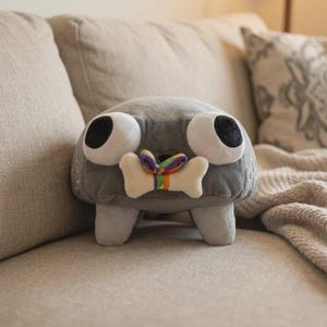 May include: A gray plush toy with large white eyes and black pupils. It has a bone-shaped detail with a rainbow ribbon. The toy is on a beige couch with decorative pillows and a blanket.