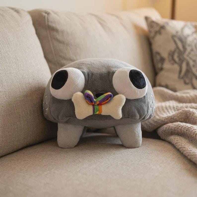 May include: A gray plush toy with large white eyes and black pupils. It has a bone-shaped detail with a rainbow ribbon. The toy is on a beige couch with decorative pillows and a blanket.
