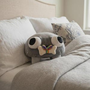 May include: A gray plush toy with large, round eyes and a bone-shaped detail with a rainbow ribbon. The toy has small legs and is resting on a bed with white pillows and a gray blanket. The toy is a whimsical, decorative item.