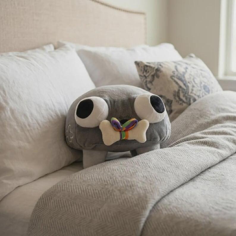 May include: A gray plush toy with large, round eyes and a bone-shaped detail with a rainbow ribbon. The toy has small legs and is resting on a bed with white pillows and a gray blanket. The toy is a whimsical, decorative item.