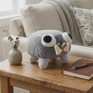 May include: A gray plush toy with large white-rimmed eyes and a rainbow-colored bow tie. The toy has four white legs and a four-leaf clover design on its side. It is sitting on a wooden table.