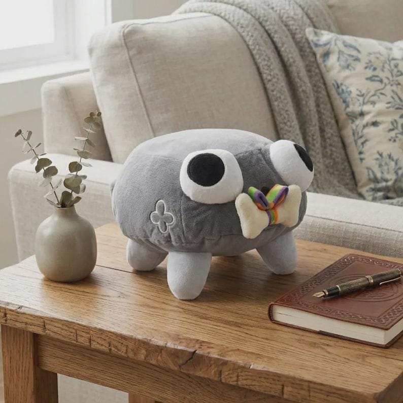 May include: A gray plush toy with large white-rimmed eyes and a rainbow-colored bow tie. The toy has four white legs and a four-leaf clover design on its side. It is sitting on a wooden table.