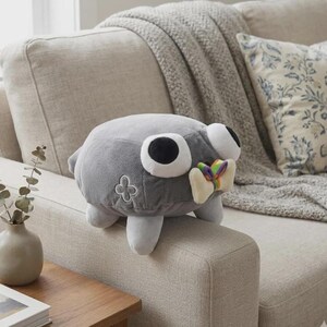May include: A gray plush toy shaped like a creature with large, round eyes and a colorful bow tie. The toy has white feet and a four-leaf clover design on its side. It is sitting on a beige couch.