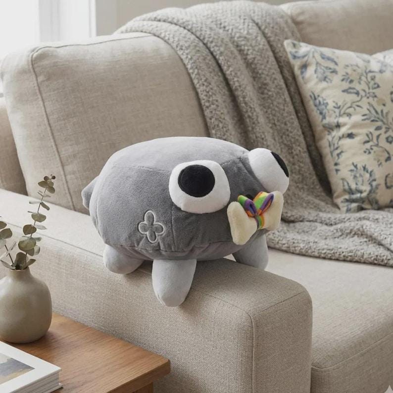 May include: A gray plush toy shaped like a creature with large, round eyes and a colorful bow tie. The toy has white feet and a four-leaf clover design on its side. It is sitting on a beige couch.