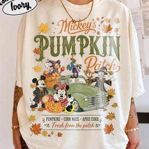 May include: An ivory t-shirt featuring a graphic of Mickey Mouse and friends in a pumpkin patch with a green vintage truck. The text reads Mickey's PUMPKIN Patch, PUMPKINS - CORN MAZE - APPLE CIDER, and Fresh from the patch, surrounded by autumn leaves.