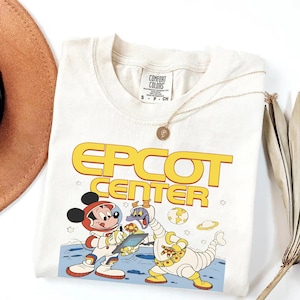 May include: Cream-colored t-shirt featuring the words "EPCOT CENTER" in yellow. The graphic shows Mickey Mouse and a purple alien in space suits. A gold necklace and a brown hat are also visible.