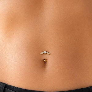 May include: A gold-toned navel piercing jewellery piece featuring a curved bar with a cluster of five clear, round-cut stones arranged in an arc shape above the belly button, complemented by a small gold ball at the bottom of the piercing.