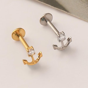 May include: Two labret body jewelry pieces featuring an anchor design. One piece is gold-toned and the other is silver-toned. Each anchor has a small, clear, round-cut gemstone set at the top center, with a flat circular backing disc.