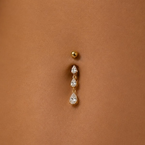 Peut inclure: Un bijou de piercing pour nombril doré composé d'une bille supérieure lisse et d'une chaîne verticale pendante. La chaîne est ornée de trois pierres transparentes en forme de poire à facettes, de taille croissante vers le bas.