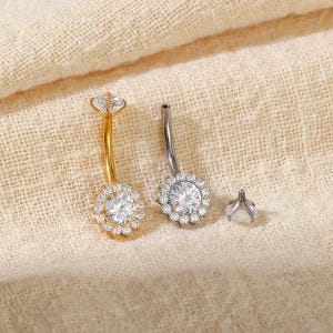 Puede incluir: Dos piercings de ombligo curvos, uno dorado y otro plateado, cada uno con una gran piedra redonda transparente central rodeada por un halo de piedras transparentes más pequeñas. Se incluye una pieza superior de rosca plateada a juego.