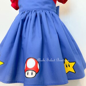 May include: A blue dress with red short sleeves and a yellow button. The dress has a mushroom and a star graphic on the skirt. The text "Akabi Select Boutique" is printed on the skirt.