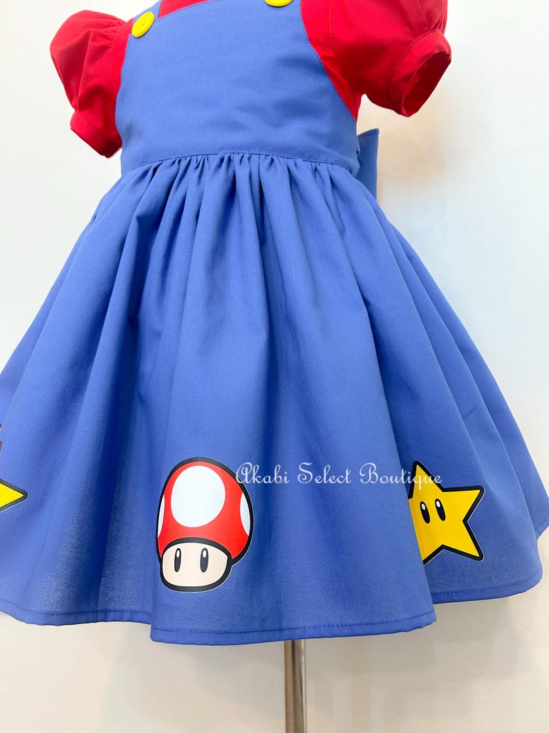 May include: A blue dress with red short sleeves and a yellow button. The dress has a mushroom and a star graphic on the skirt. The text "Akabi Select Boutique" is printed on the skirt.