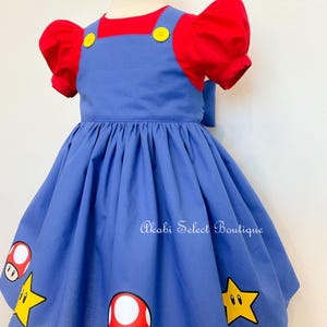 May include: A blue overall dress with red short sleeves and yellow buttons. The dress has a white and red mushroom and a yellow star graphic print. The dress is on a white mannequin.