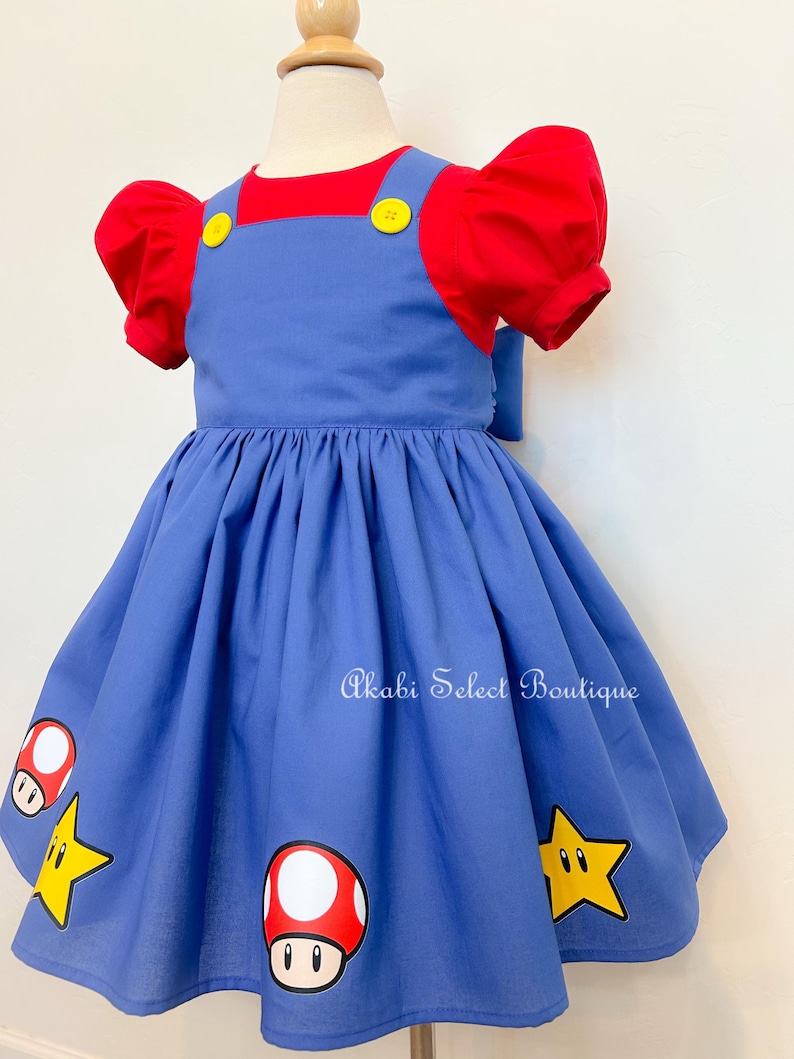 May include: A blue overall dress with red short sleeves and yellow buttons. The dress has a white and red mushroom and a yellow star graphic print. The dress is on a white mannequin.
