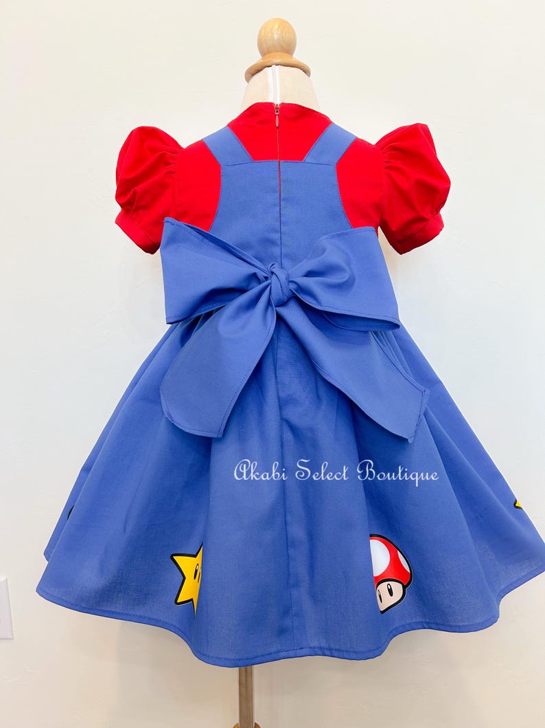 May include: A blue dress with a red top and a large bow in the back. The dress has a star and mushroom design. The text "Akabi Select Boutique" is printed on the dress.