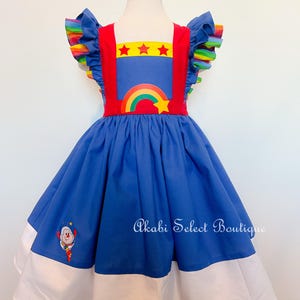 Puede incluir: Un vestido azul y blanco con detalles de arcoíris y estrellas. El vestido tiene tirantes rojos y un corpiño rojo con un arcoíris y una estrella. El vestido tiene un volante blanco alrededor del dobladillo y un volante blanco alrededor de las mangas. El vestido tiene un payaso bordado en la parte delantera. El vestido está en un maniquí blanco.