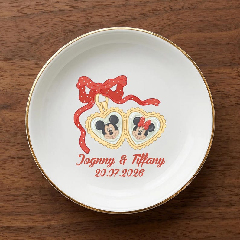 Op de afbeelding: Een witte keramische plaat met een gouden rand. Het toont Mickey en Minnie Mouse in hartvormige frames, een rode strik met witte stippen en de namen "Jognny & Tiffany" met de datum "20.07.2026".