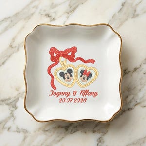 Op de afbeelding: Wit keramisch schaaltje met een gouden rand en geschulpte randen. Voorzien van een rode strik en hartvormige hangers met Mickey en Minnie Mouse. De tekst "Jognny & Tiffany 20.07.2026" is rood gedrukt.