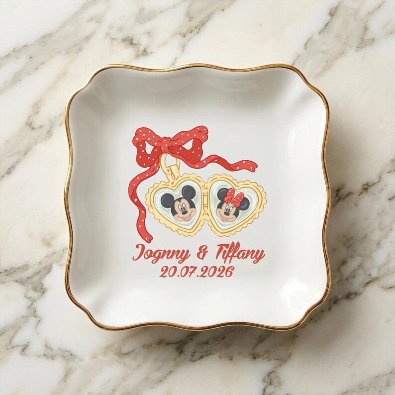 Op de afbeelding: Wit keramisch schaaltje met een gouden rand en geschulpte randen. Voorzien van een rode strik en hartvormige hangers met Mickey en Minnie Mouse. De tekst "Jognny & Tiffany 20.07.2026" is rood gedrukt.