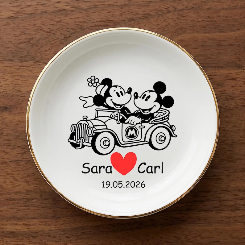 Op de afbeelding: Een witte keramische plaat met een gouden rand toont een zwarte illustratie van Mickey en Minnie Mouse in een oldtimer. De plaat bevat de namen "Sara" en "Carl", een rood hart en de datum "19.05.2026".