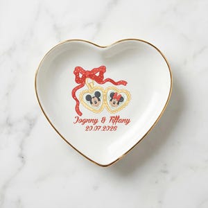 Op de afbeelding: Hartvormige witte keramische sieradenschaal met een gouden rand. De schaal heeft een rode strik en twee hartvormige medaillons met Mickey en Minnie Mouse. De namen "Johnny & Tiffany" en de datum "20.07.2026" zijn rood gedrukt.