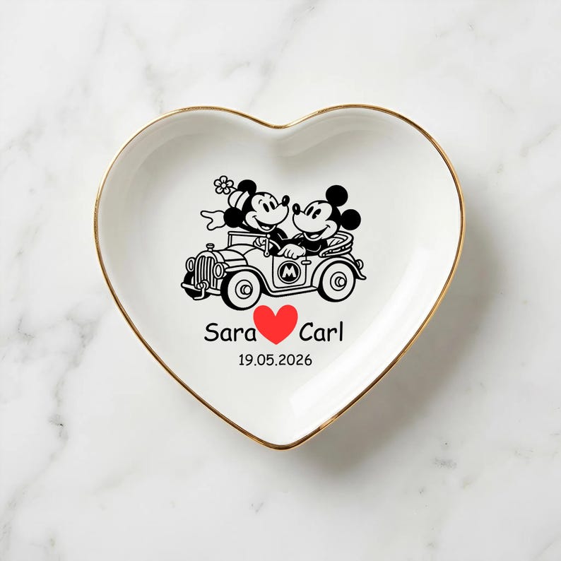 Op de afbeelding: Wit hartvormig keramisch schaaltje met een gouden rand, met een zwart-rode afbeelding van Mickey en Minnie Mouse in een oldtimer. De namen "Sara" en "Carl" staan eronder, met een rood hart en de datum "19.05.2026".
