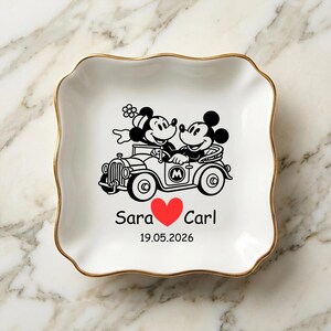 Op de afbeelding: Wit keramisch sieradenschaaltje met een gouden rand. Het schaaltje heeft een zwart-rode afbeelding van Mickey en Minnie Mouse in een oldtimer, met de namen "Sara" en "Carl" en een datum eronder. Het schaaltje heeft een golvende rand.