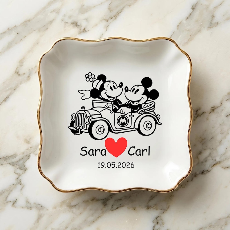 Op de afbeelding: Wit keramisch sieradenschaaltje met een gouden rand. Het schaaltje heeft een zwart-rode afbeelding van Mickey en Minnie Mouse in een oldtimer, met de namen "Sara" en "Carl" en een datum eronder. Het schaaltje heeft een golvende rand.