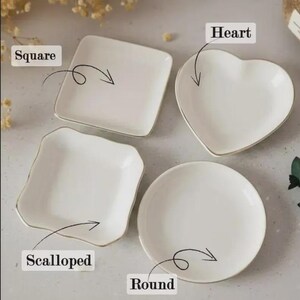 Op de afbeelding: Vier witte keramische schalen in verschillende vormen: vierkant, hart, geschulpt en rond. Elke schaal heeft een gouden rand. De vormen zijn gelabeld met de woorden "Square", "Heart", "Scalloped" en "Round".
