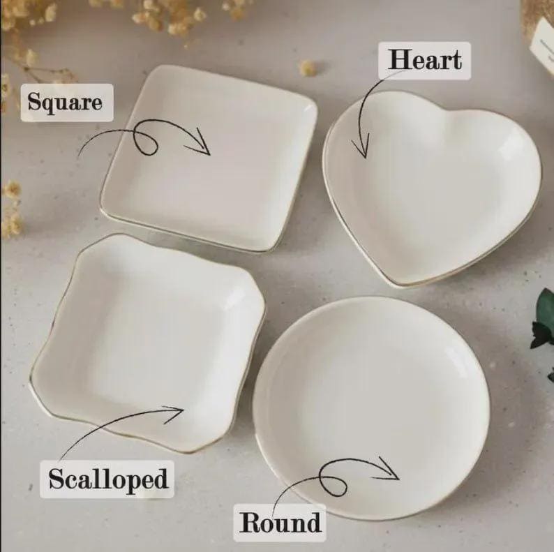Op de afbeelding: Vier witte keramische schalen in verschillende vormen: vierkant, hart, geschulpt en rond. Elke schaal heeft een gouden rand. De vormen zijn gelabeld met de woorden "Square", "Heart", "Scalloped" en "Round".
