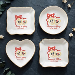 Op de afbeelding: Vier witte decoratieve schaaltjes met gouden rand, elk met een Mickey en Minnie Mouse ontwerp met een rode strik en de tekst "Jognny & Tiffany 20.07.2026". De schaaltjes hebben vierkante, ronde, hart- en geschulpte vierkante vormen.