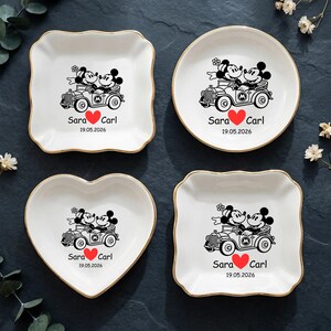 Op de afbeelding: Vier witte keramische sieradenschaaltjes met gouden randen. Elk schaaltje heeft een zwarte illustratie van Mickey en Minnie Mouse in een oldtimer, met de namen "Sara" en "Carl", een rood hart en de datum "19.05.2026". De schaaltjes zijn vierkant, rond, hartvormig en vierkant met geschulpte rand.