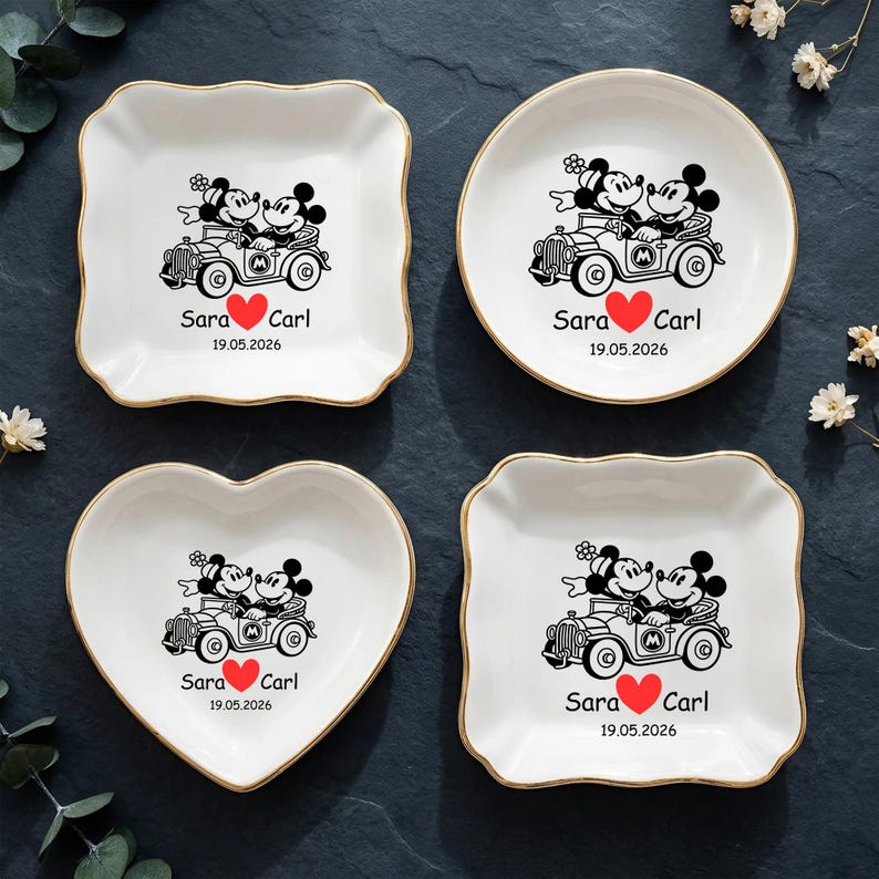 Op de afbeelding: Vier witte keramische sieradenschaaltjes met gouden randen. Elk schaaltje heeft een zwarte illustratie van Mickey en Minnie Mouse in een oldtimer, met de namen "Sara" en "Carl", een rood hart en de datum "19.05.2026". De schaaltjes zijn vierkant, rond, hartvormig en vierkant met geschulpte rand.